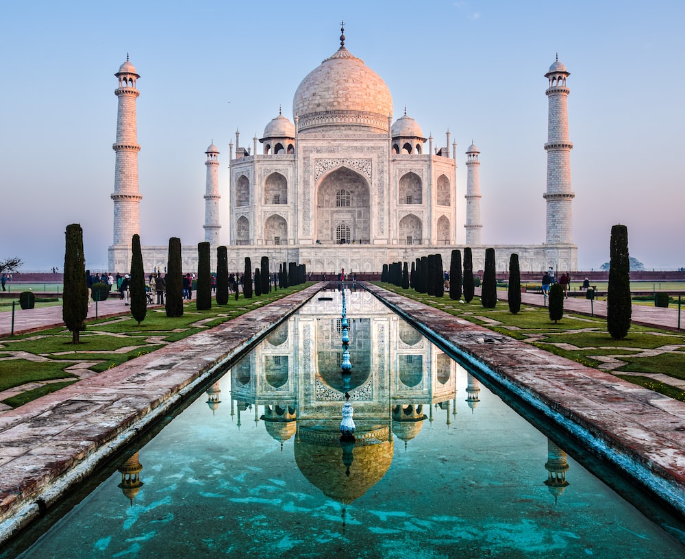 The image captures the architectural masterpiece of the Taj Mahal in Agra at sunrise, its white marble dome and minarets reflecting in a long water pool lined with shrubs and trees.