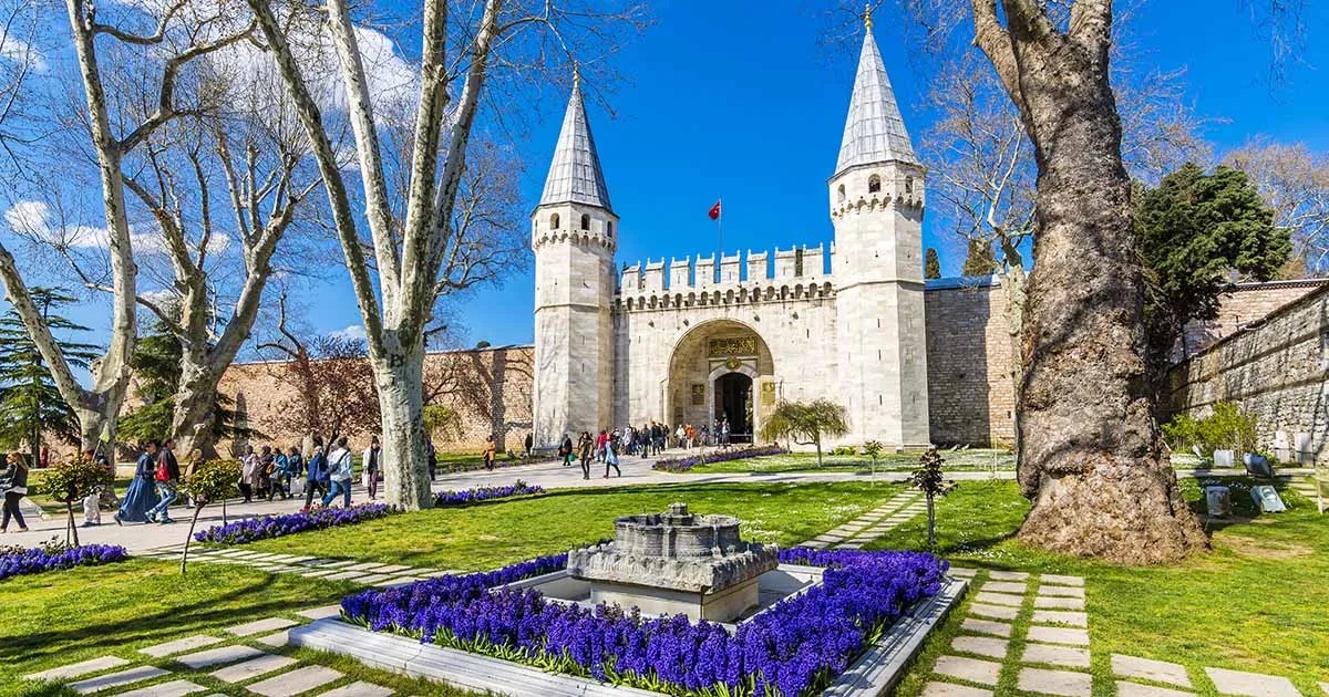 Entrance of Topkapi Palace Museum in Istanbul, Turkey, featuring historic Ottoman architecture, stone towers, gardens with purple flowers, and visitors walking under clear blue sky.