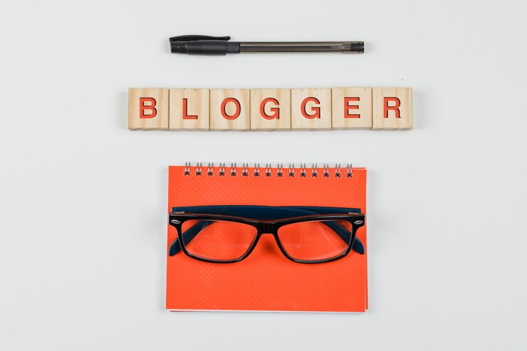 A pen, "blogger" spelled with wooden blocks, and a spiral notebook with a pair of glasses on top