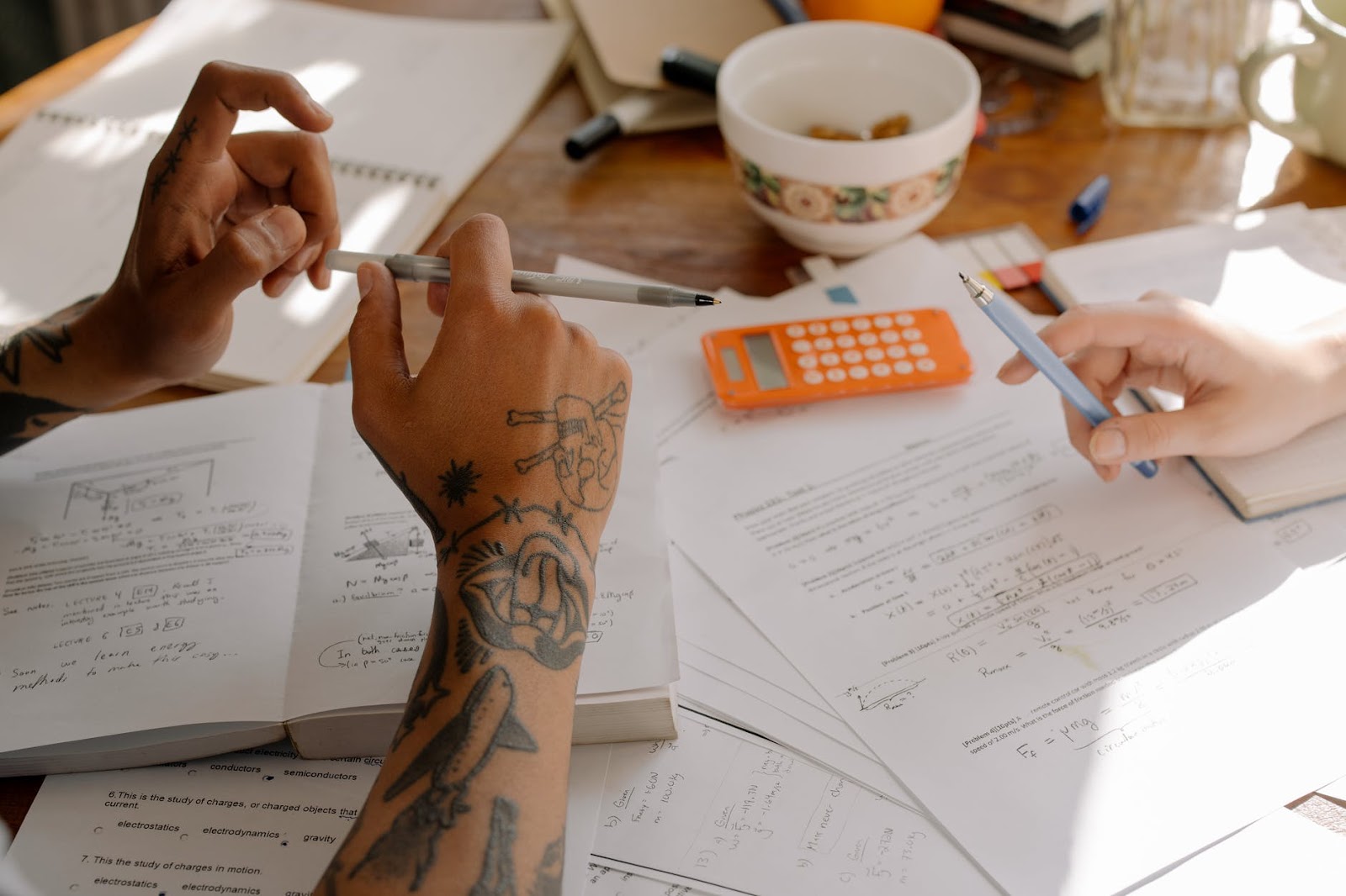 Two people are sitting at a table filled with papers, notes, and books, engaged in a discussion or study session. They are holding pens and pointing to different sections of the documents. A calculator and a bowl are also visible on the table as they prepare for the National Aptitude Test in Architecture.