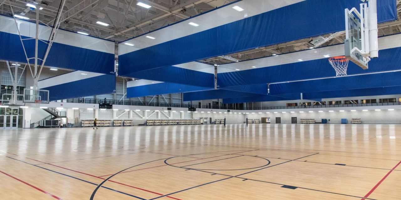 Un gymnase intérieur spacieux avec un sol en bois poli, plusieurs paniers de basket et de grands rideaux de séparation rétractables bleus et blancs suspendus au plafond.