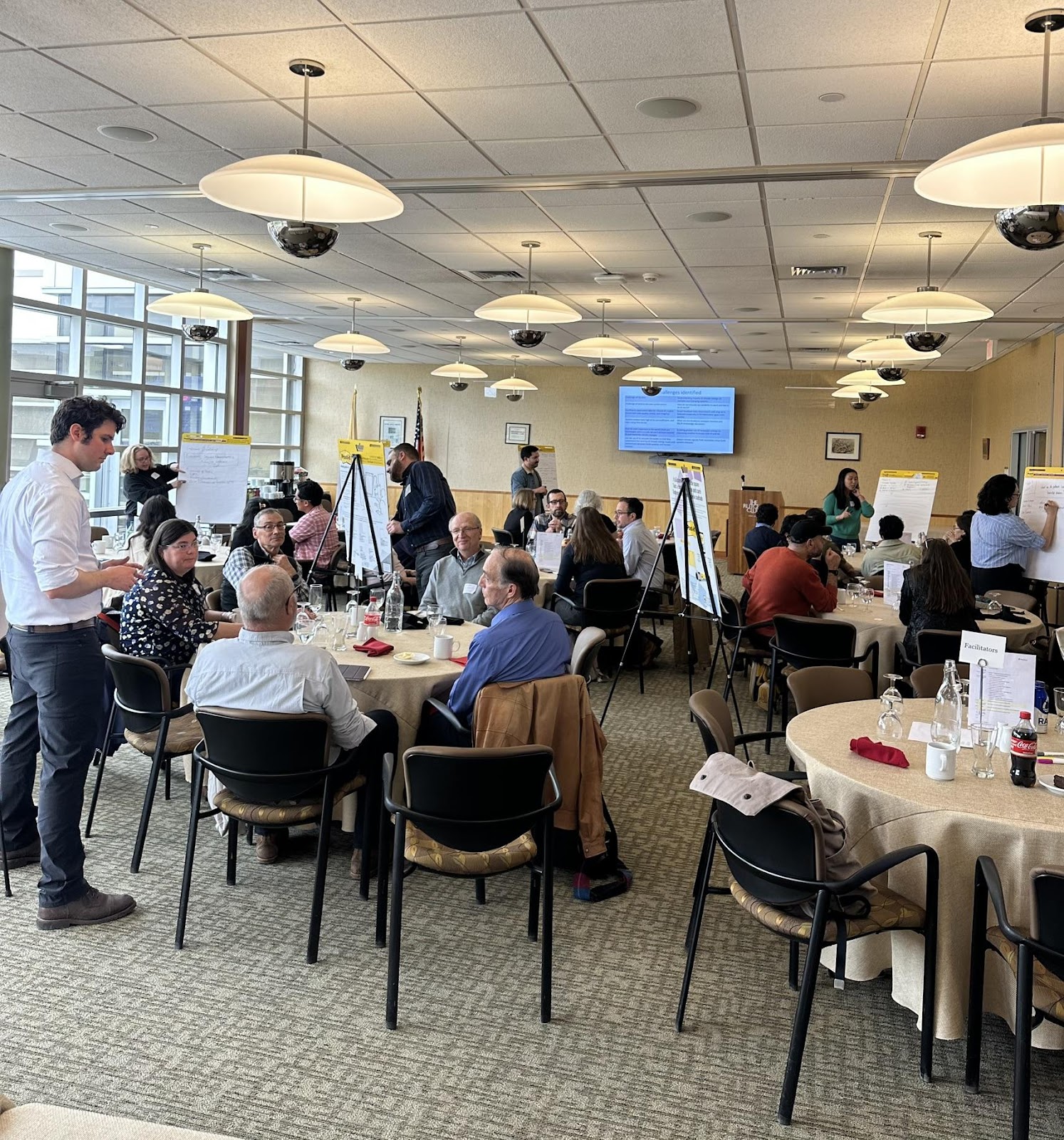 A networking session between participants in a cafe style setting with circular tables, chairs, and large easel notepads