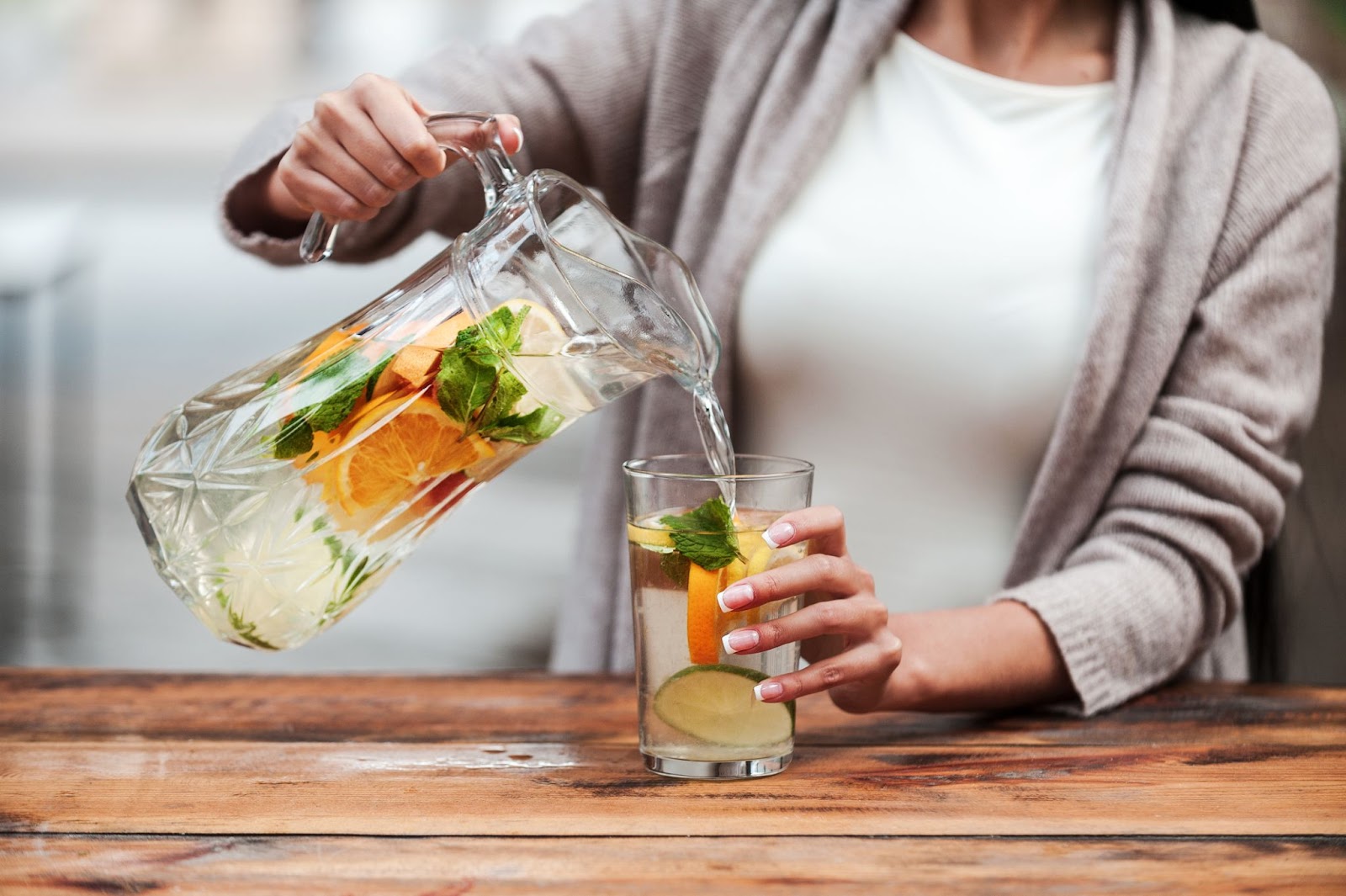 A cropped shot of a woman pouring orange, cucumber, and mint-infused water from a glass pitcher into a glass on a wooden table.