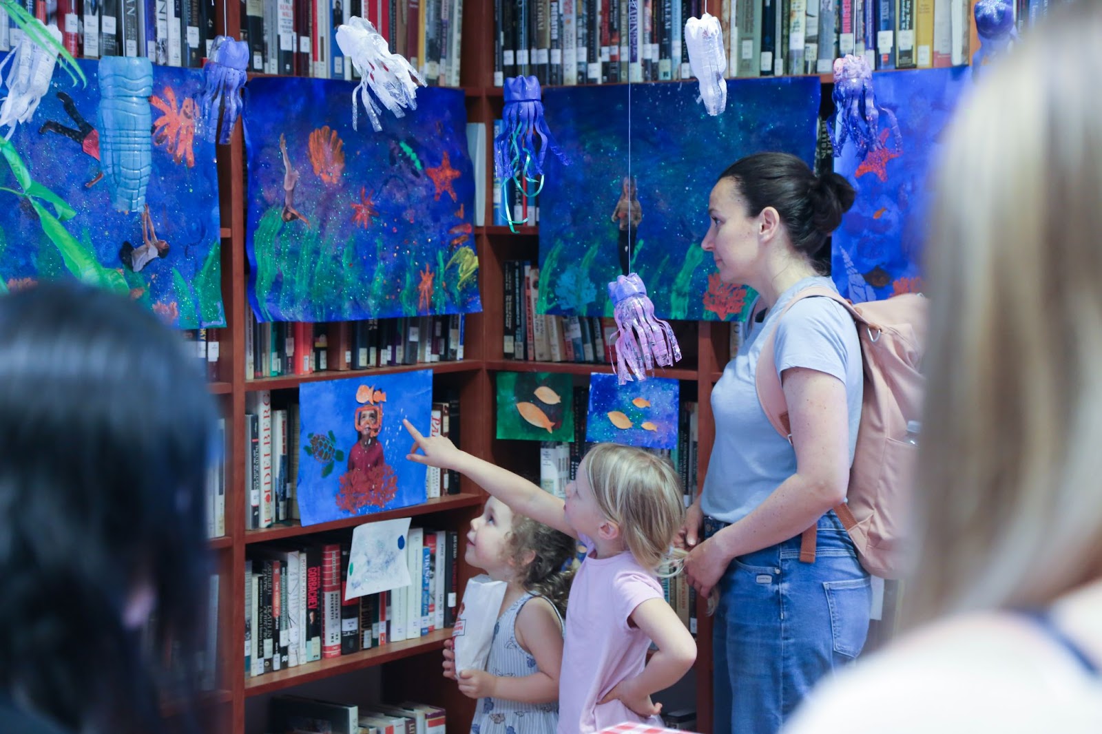 Two young girls pointing at a watercolour painting and collage, while an adult woman stands behind them. More such paintings are displayed on bookshelves around the girls. 