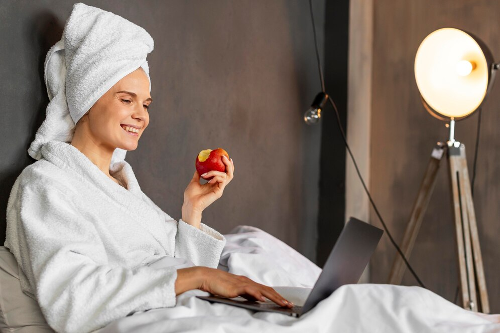 A woman relaxing in a hotel room, working on a laptop and eating an apple.