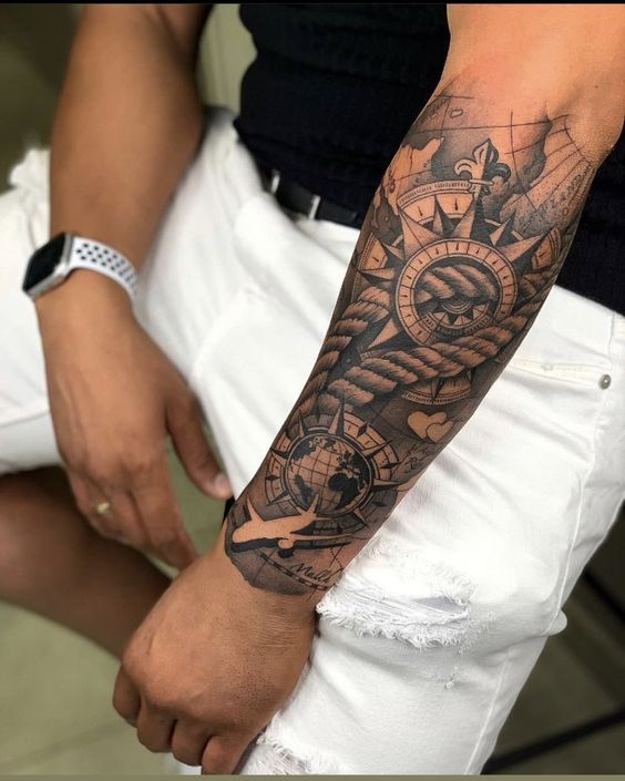 Realistic forearm tattoo featuring a compass, ship anchor, and world map design on a person wearing white pants and a smartwatch.