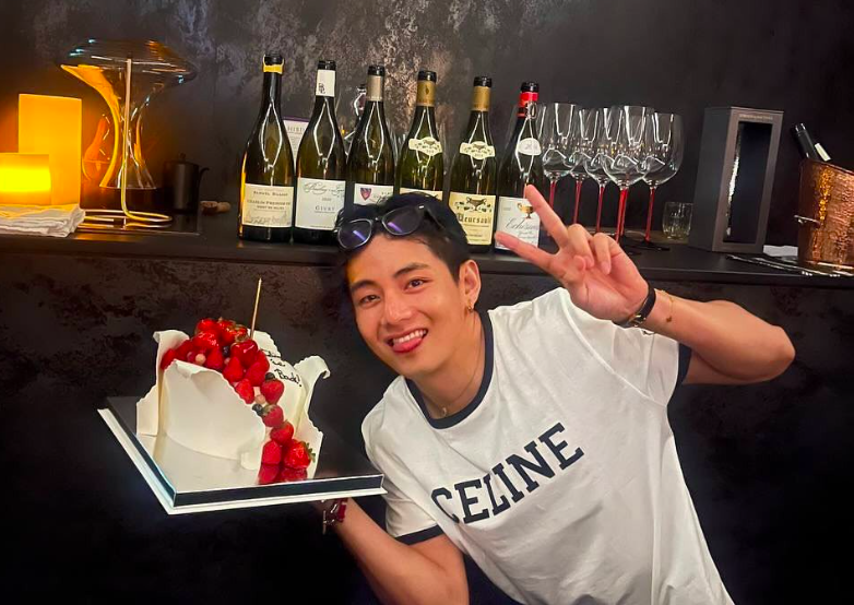 This contains an image of BTS V holding a cake and making peace sign with his left hand