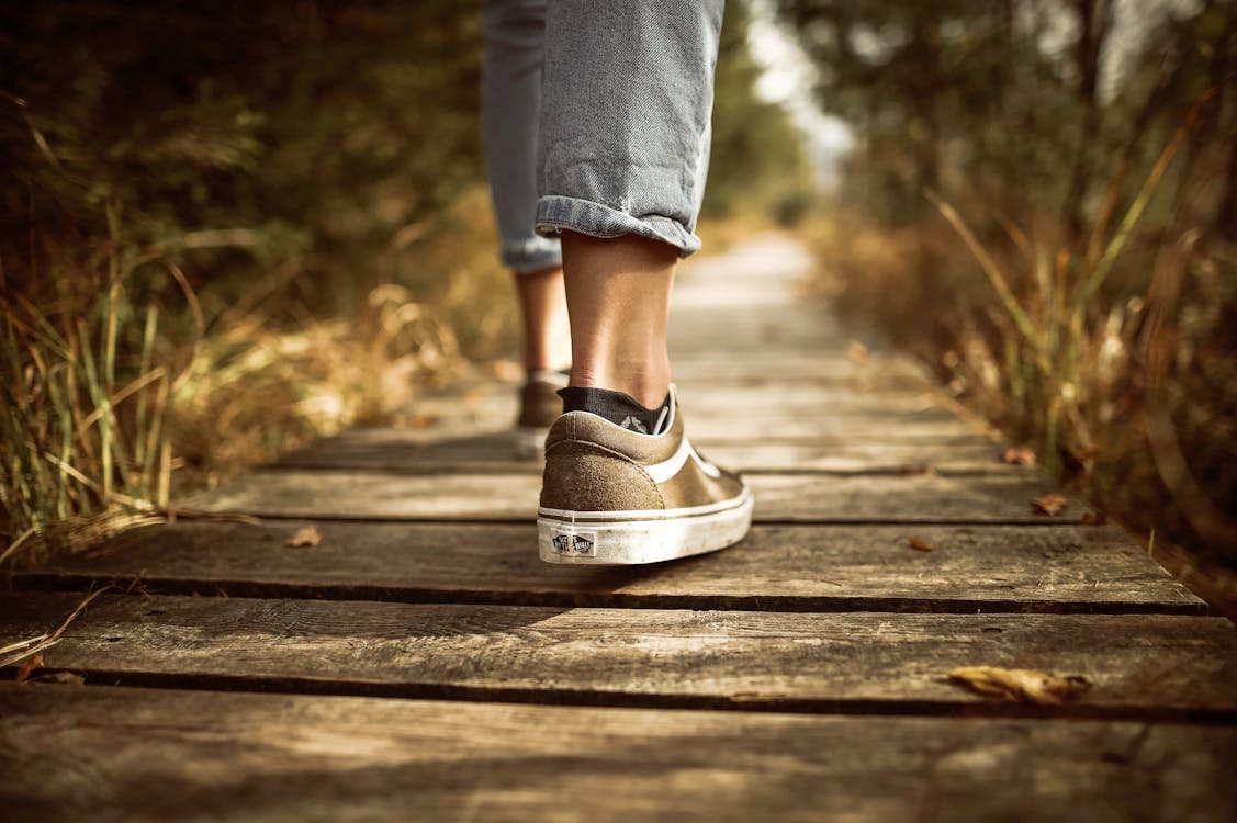 A photograph of two legs with feet wearing brown trainers and black trainer socks, and pale blue ankle length jeans. The person is walking on wood boards that are a path beside late summer grasses and trees.