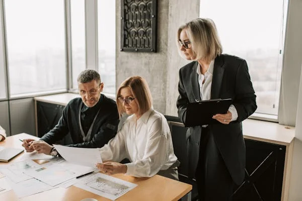 An experienced woman supervisor in a black dress stands confidently, holding a pad while overseeing her two colleagues, guiding them with their duties