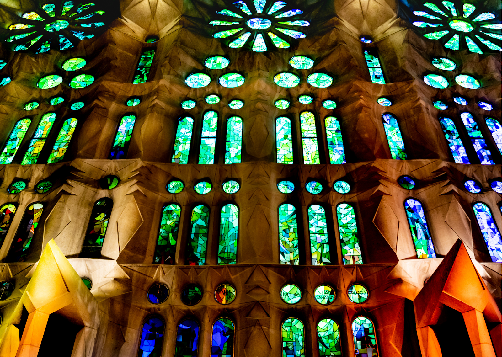 This image captures an unspoken rule for tourists in Spain: appreciating the cultural and spiritual significance of local landmarks. It showcases the stunning stained glass windows of a Spanish cathedral, which are illuminated by vibrant hues of green, blue, and yellow. The intricate designs and geometric patterns create a kaleidoscope of colors that dance across the stone interior, highlighting the grandeur and spiritual awe of the architecture. The vivid lighting emphasizes the craftsmanship of the stained glass, drawing the viewer's gaze upward and inviting a deeper appreciation for Spain's sacred spaces and cultural heritage.