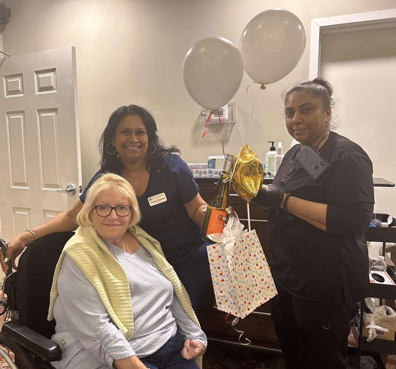 A woman smiling in a wheelchair with caregivers with balloons in her assisted living apartment