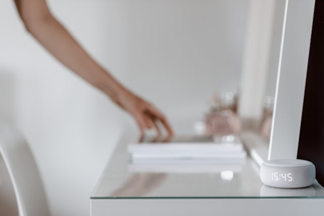 Assistente Alexa na cor branca mostrando 15:45 em uma mesa de vidro, enquanto uma mão ao fundo interage com objetos.