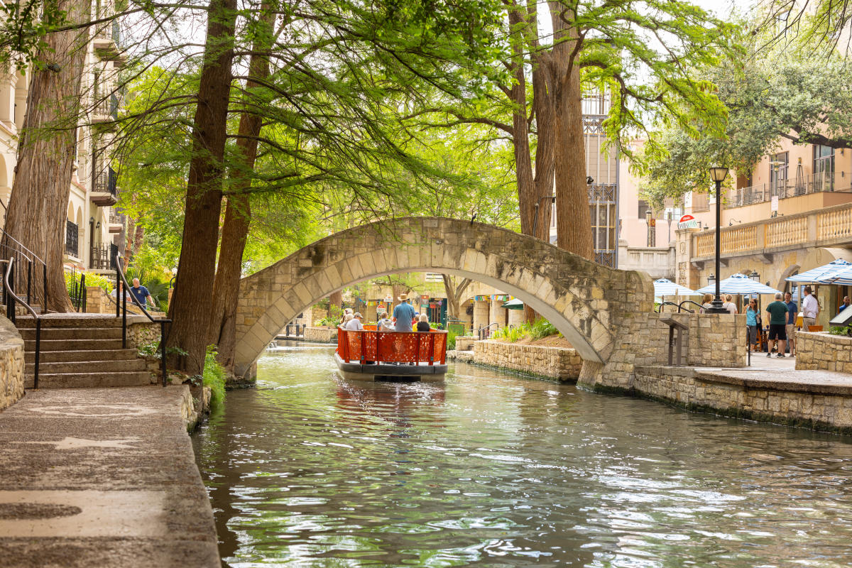 The San Antonio River Walk with a stone arch bridge, a red boat with passengers, and people walking along the riverside path.