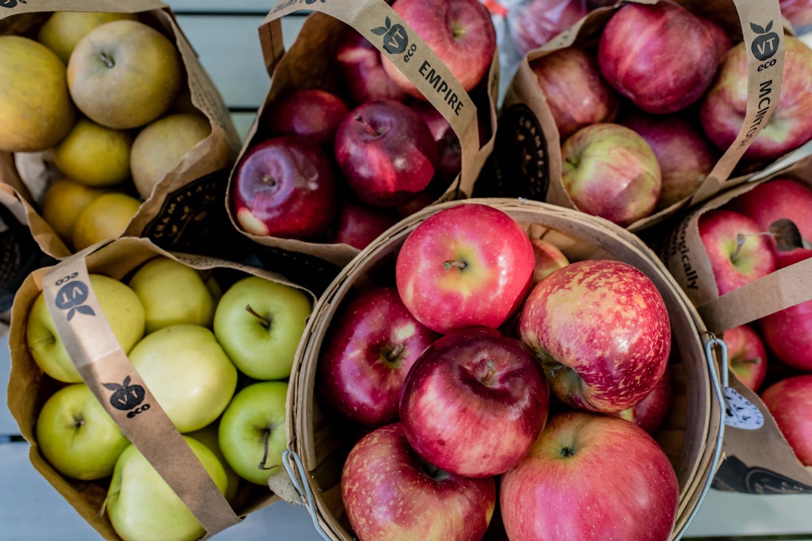 Assorted apples in paper bags and a basket, including green and red varieties like Empire and McIntosh, displayed for sale.