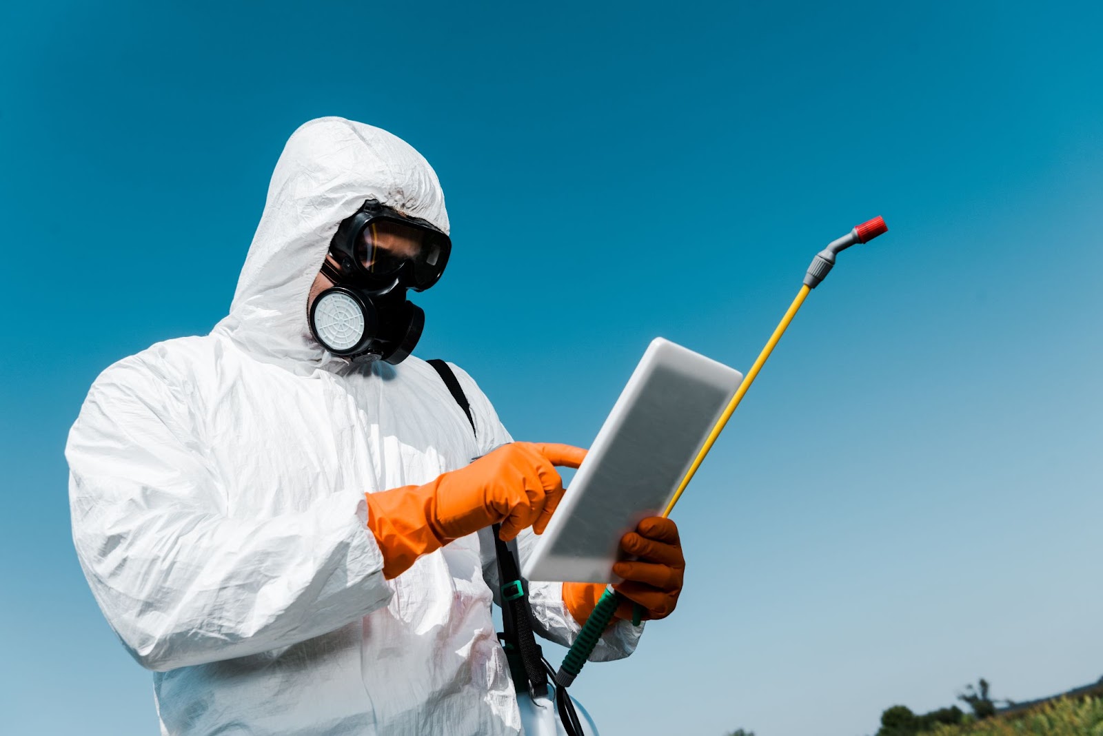 Professional pest control worker in protective gear using a tablet for treatment guidance in an outdoor area.