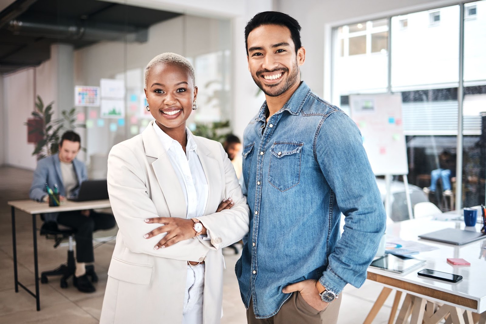 Man and a woman smiling with people and desks in the background. 