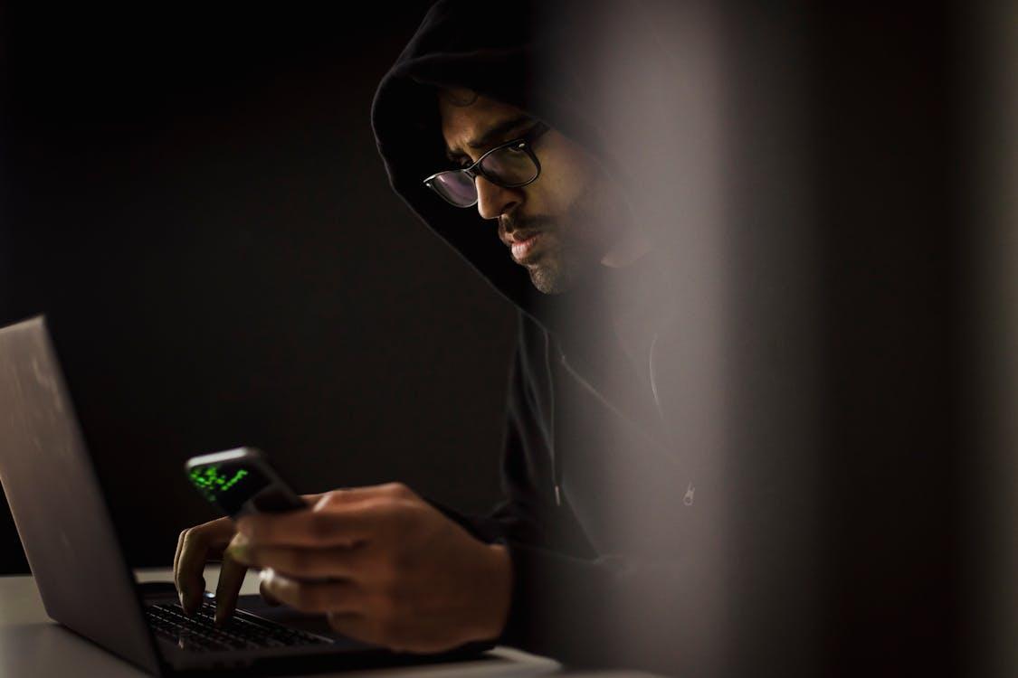 Free Man in hoodie using laptop and smartphone in dark room, focused and concentrated. Stock Photo