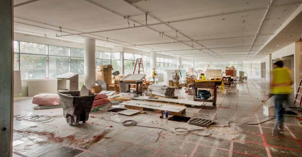(A spacious indoor construction site filled with tools, materials, and workers in safety vests, showcasing an active commercial renovation in progress.)