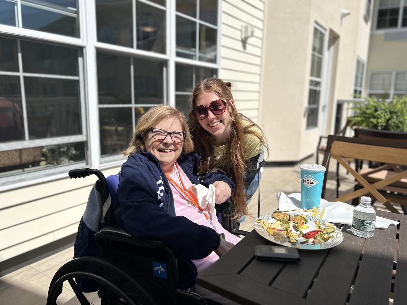 An elderly woman smiling with a caregiver while eating a meal outside