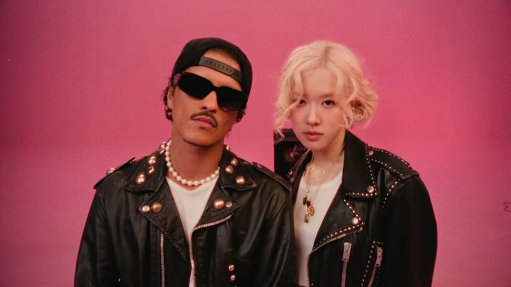 This contain BLACKPINK's Rosé and Bruno Mars standing next to each other in front of a pink background wearing black leather jackets