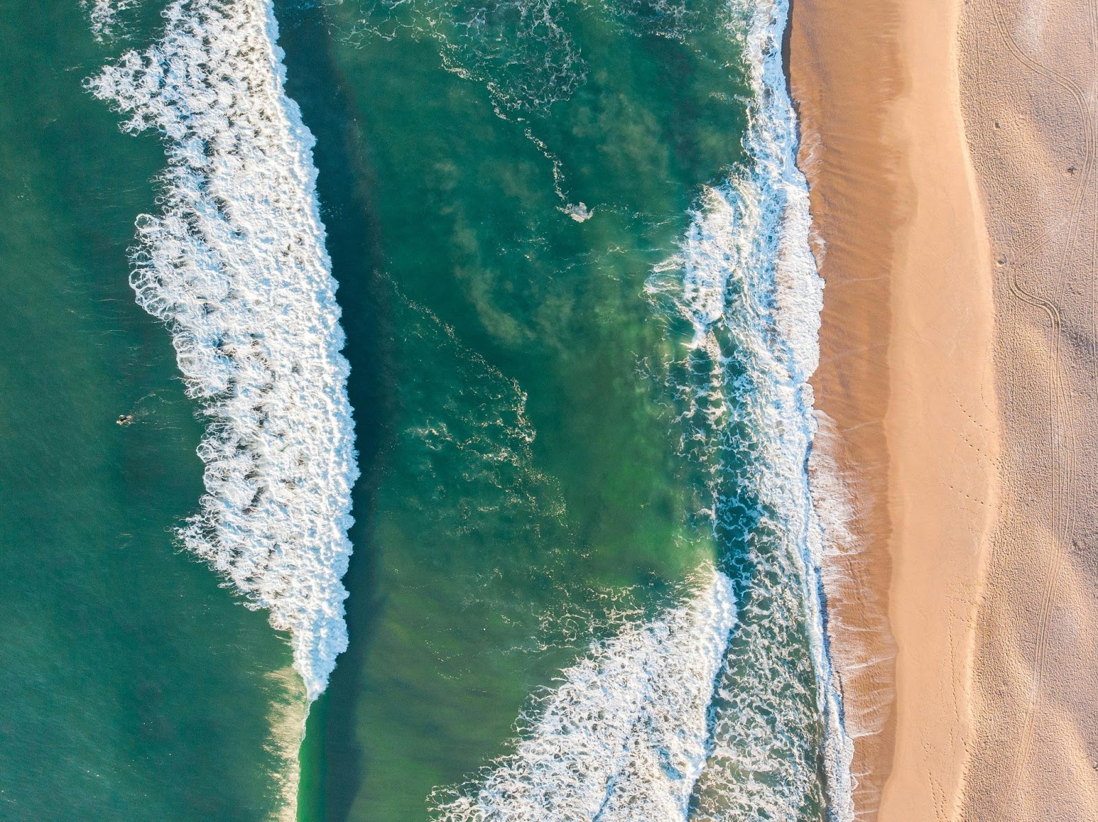 Vista aérea de uma praia, com areia fina e uniforme, com um mar verde com espuma branca das ondas.
