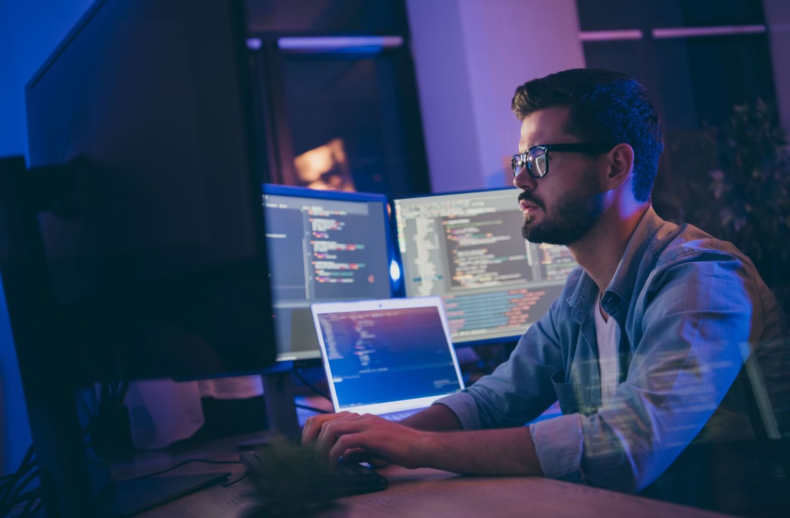 A tech worker wearing computer glasses typing on a keyboard, surrounded by multiple monitors full of code