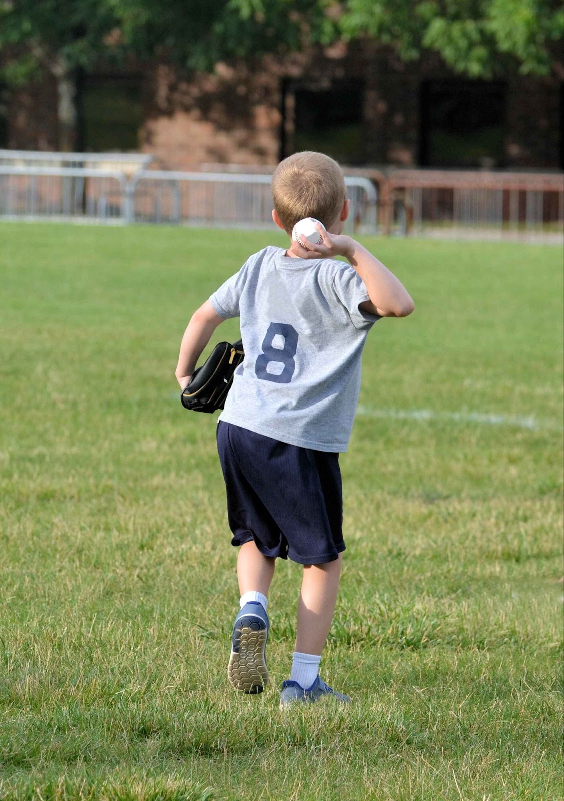 Image from behind of a young boy standing in a field throwing a baseball.
