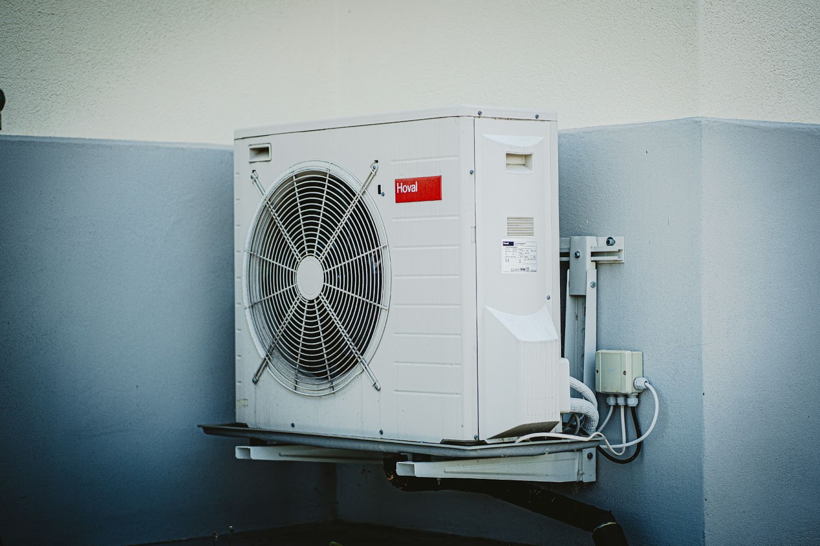 A close-up of a wall-mounted HVAC unit with a visible fan and electrical connections against a white and blue exterior wall