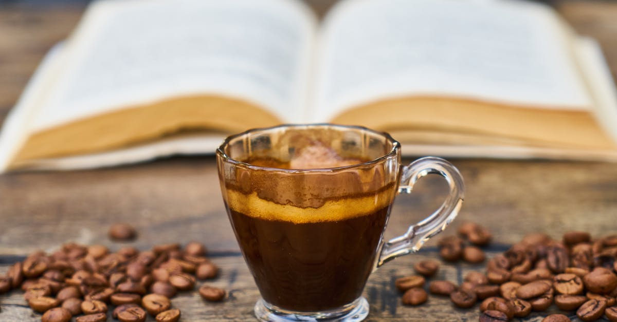 A cup of espresso with a layer of crema sits on a wooden table, surrounded by coffee beans. An open book is blurred in the background.