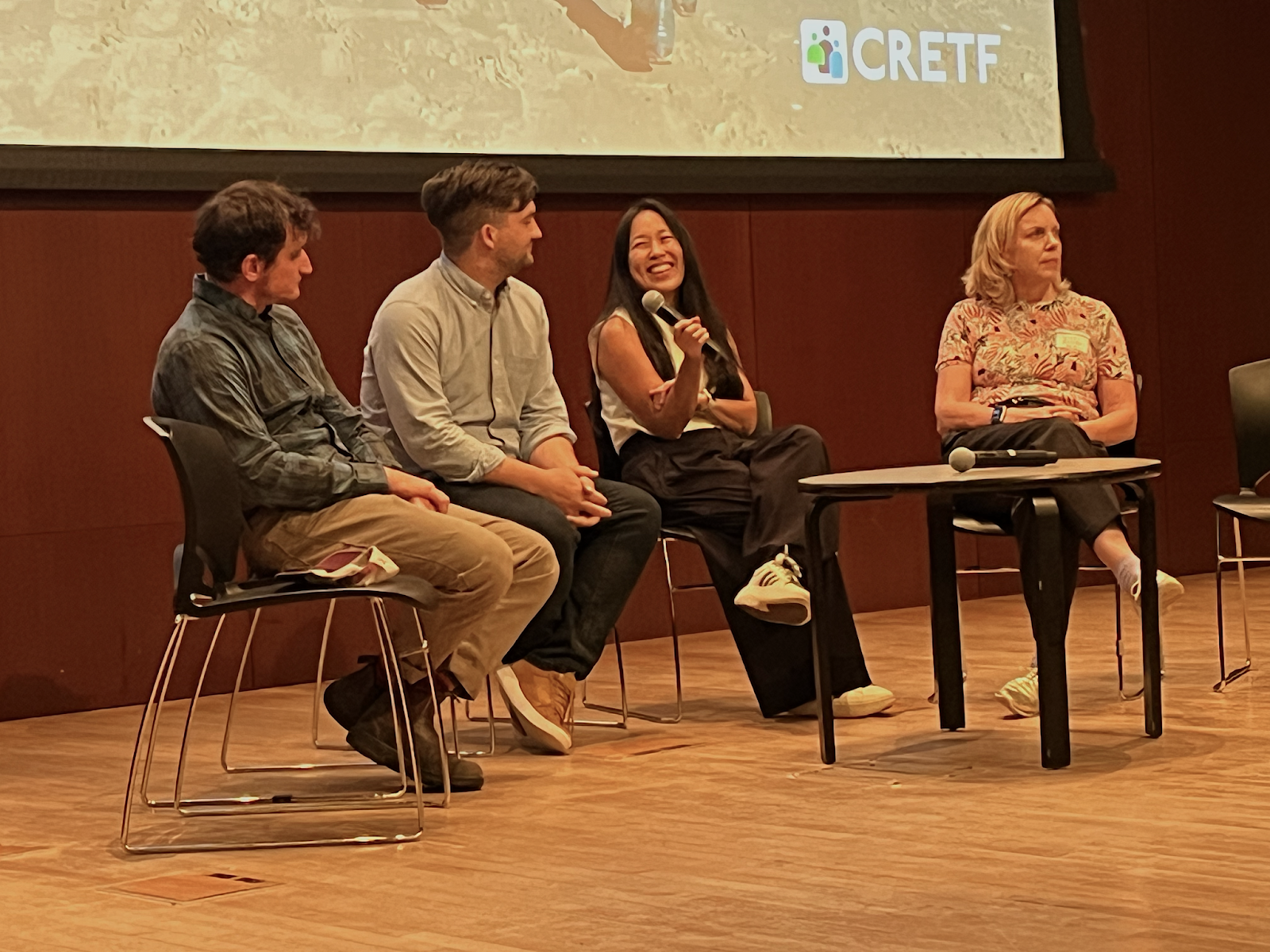 A group of people sit in a semi-circle in chairs, one person is holding a microphone.