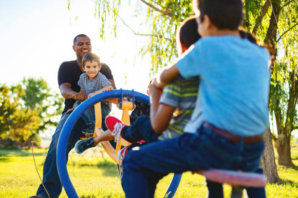 Father playing with children at a playground while other parents watch nearby, representing healthy co-parenting activities and shared parenting responsibilities in a community setting.