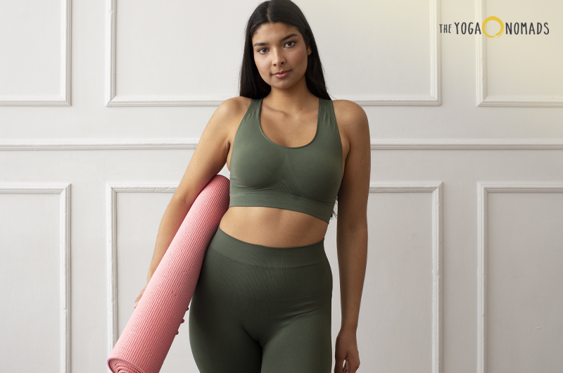 An individual is dressed in green athletic wear, consisting of a sports bra and high-waisted leggings, holding a pink yoga mat. The background features a white paneled wall, and there is a logo in the top right corner that reads "THE YOGA NOMADS".