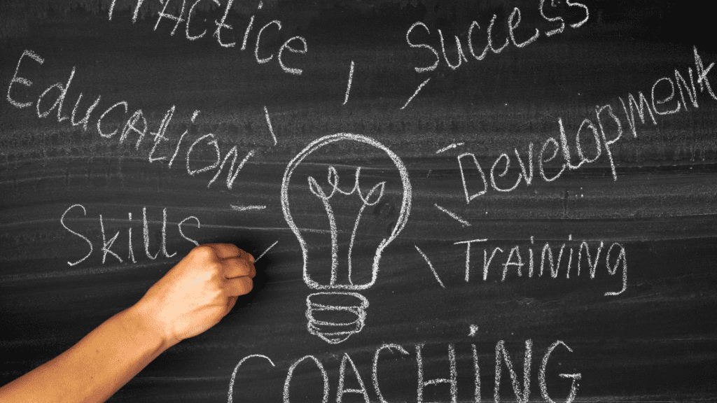 A hand drawing a lightbulb on a chalkboard with words like Coaching, Success, Development, and Skills written around it, symbolizing personal and professional growth.