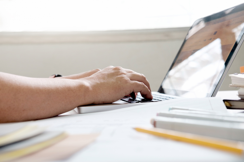 Hands typing on a laptop keyboard, perhaps searching for study tips. The laptop sits on a white desk alongside various books and papers, with a pencil visible.