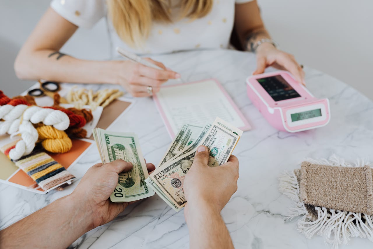 Two individuals managing cash flows, with one person counting dollar bills and the other making notes while a card payment terminal sits nearby on a table.