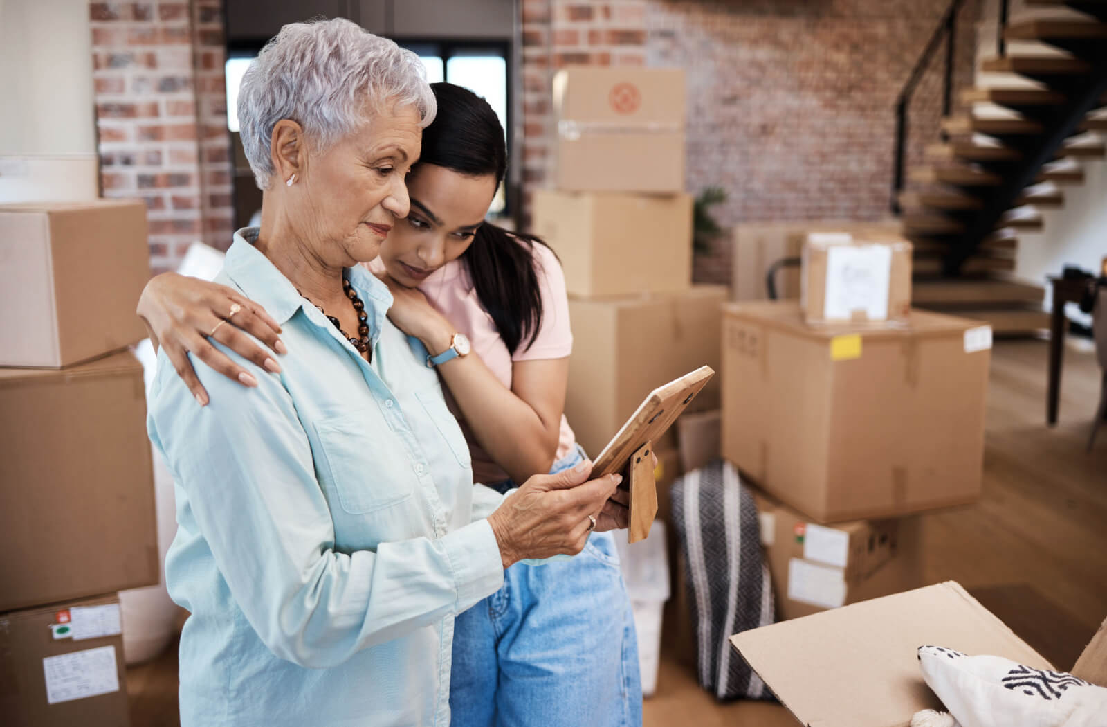 A young adult hugging their grandparent looking at a picture, surrounded by moving boxes and helping them move to assisted living.