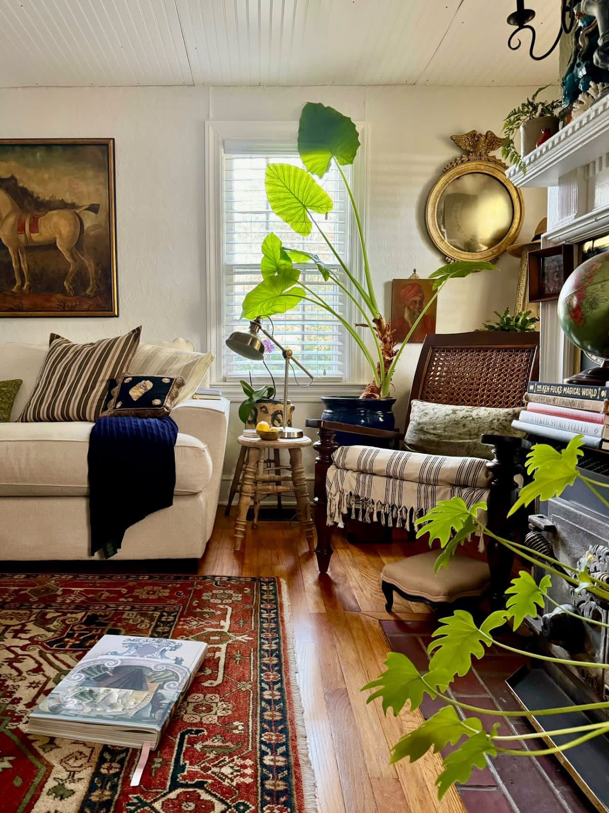 A corner in the living room next to the fireplace decorated with a large horse painting and a federal style gilted mirror hung on the wall among many other items such as white sofa, persian rug, indoor plants, etc.