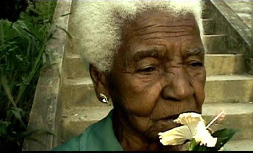 Mulher negra, idosa, com cabelo crespo branco, usando um brinco pequeno dourado. Ela veste uma blusa na cor verde e próximo à boca possui uma flor de hibisco branca. Em segundo plano, há uma escadaria.