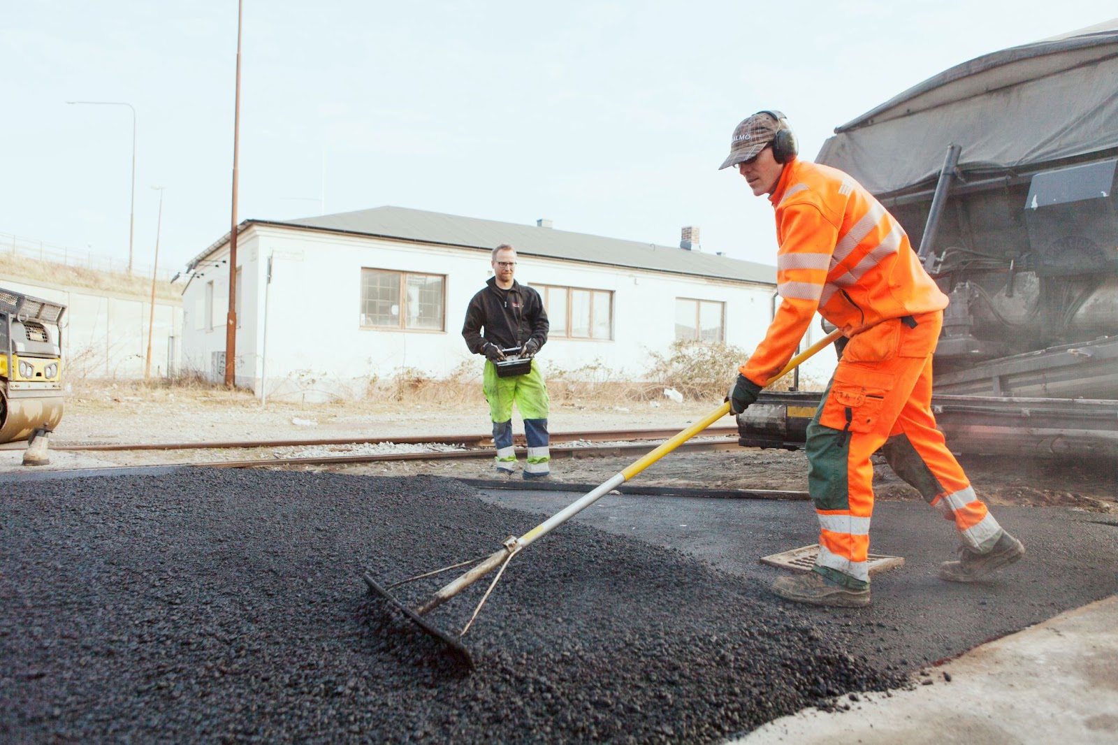 A full-length shot of a manual worker in an orange safety suit, spreading asphalt on the street with a rake as a colleague observes nearby.