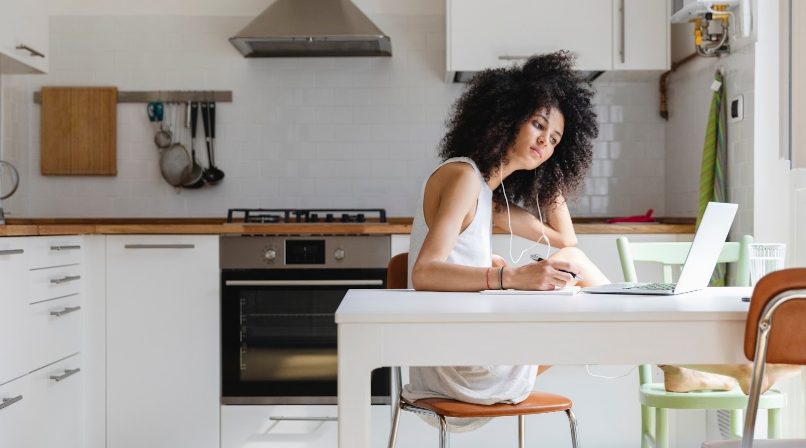 Une femme aux cheveux bouclés, portant des écouteurs, travaille sur un ordinateur portable et prend des notes à une table de cuisine.