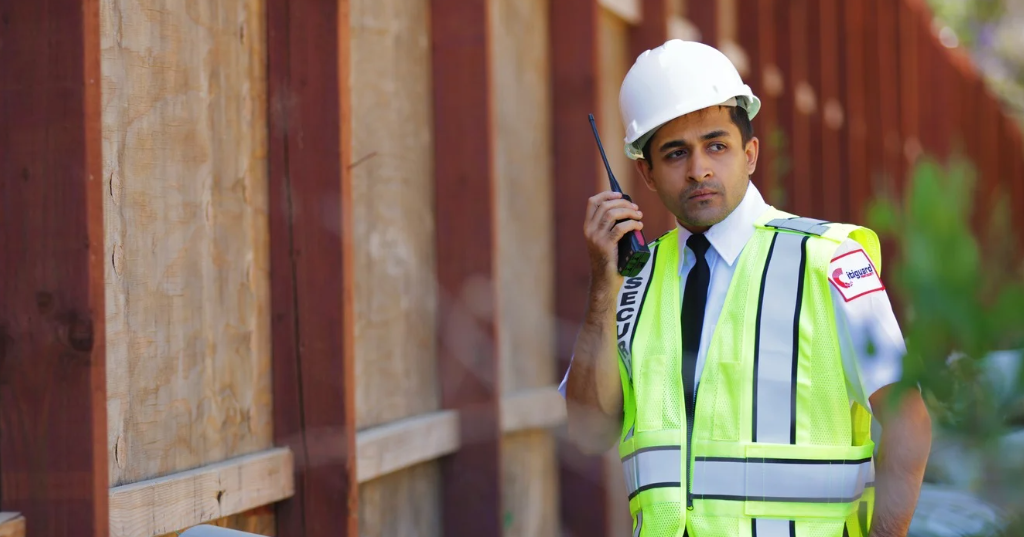 (A construction site security guard in a hard hat and reflective vest communicates via two-way radio while monitoring the perimeter of a job site.)