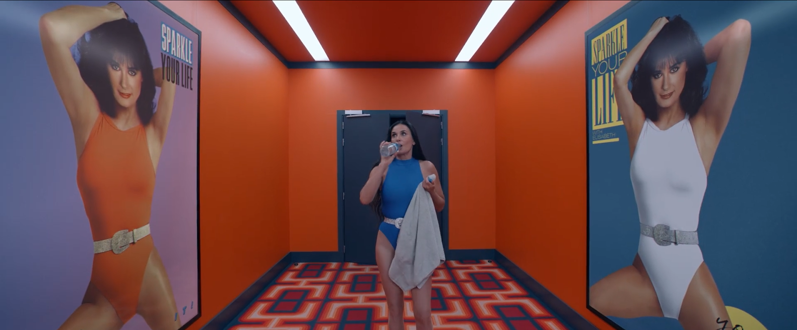 Mujer en traje de baño azul bebiendo agua mientras camina por un pasillo retro de colores naranjas y azules, con carteles en la pared que muestran imágenes de otra mujer en traje de baño y la frase 'Sparkle Your Life'. El ambiente evoca una estética de gimnasio o centro de ejercicios de los años 80.