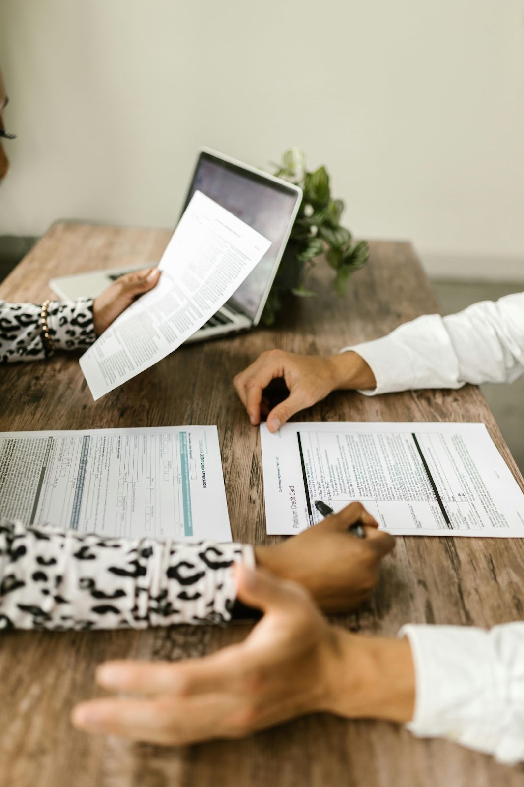 Two people review documents at a wooden table, with a laptop and plant in the background.