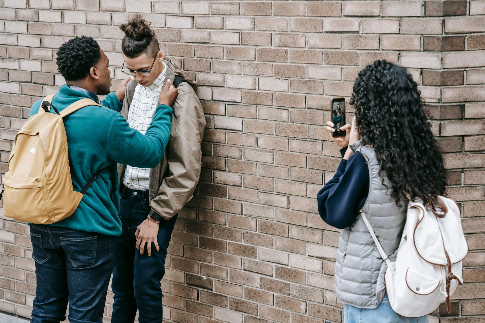 One student bullies another, pinning him to the wall, as another student records the incident with her phone