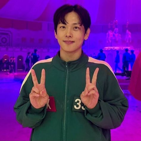 This contains an image of Squid Game 2 star Im Siwan's making the peace sign in front of a purple lit room with other people