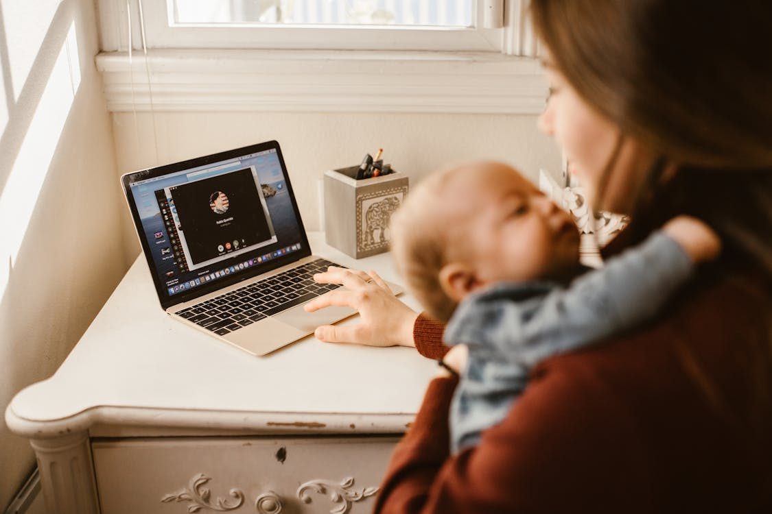 Woman holding a baby while making a video call on a laptop at a small desk, multitasking in a cozy home setting.