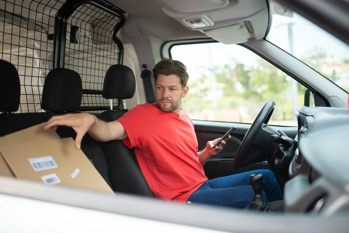 A man sitting in a van holding a phone about to deliver a parcel.