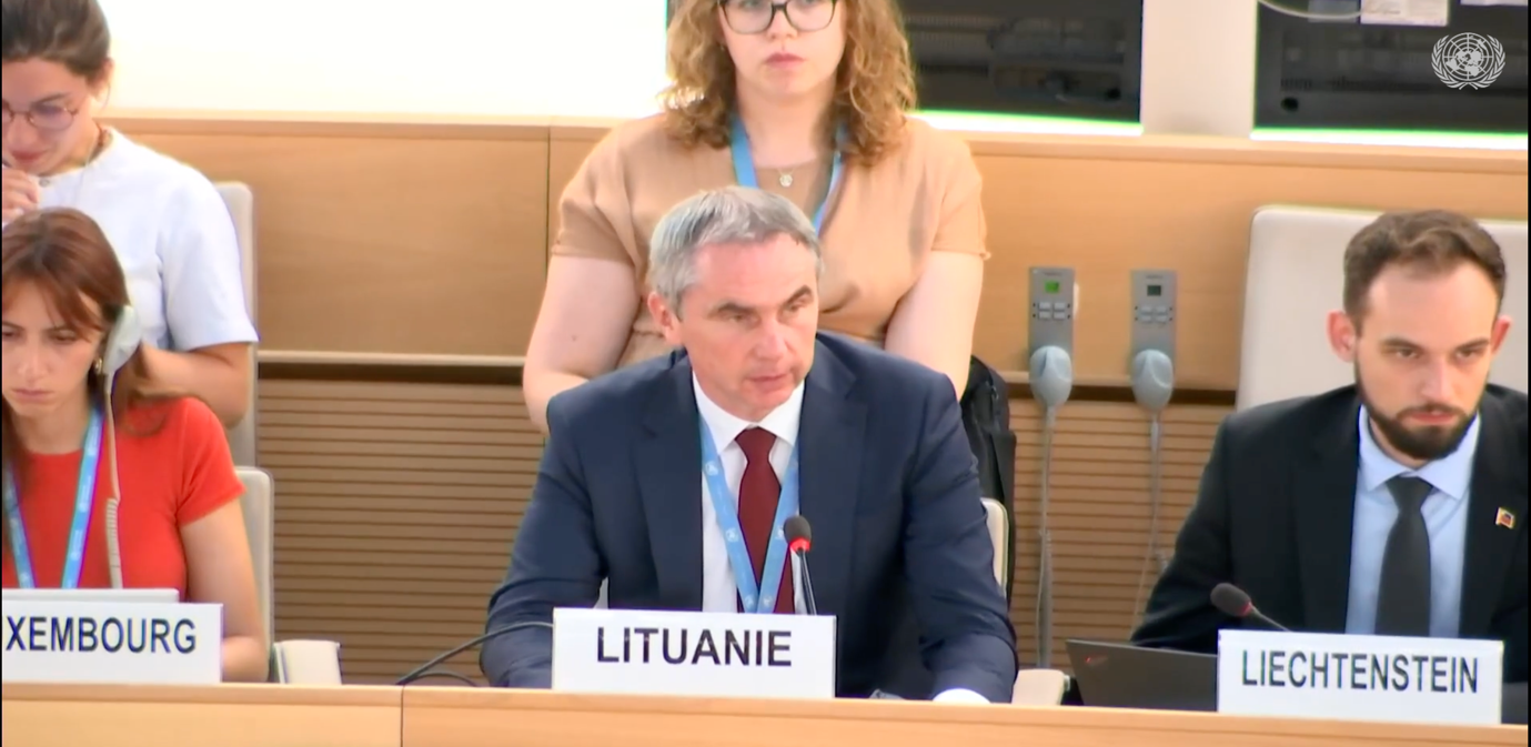 A person in a suit sitting at a podium with a person behind him

Description automatically generated