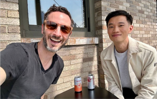 Two men sitting at a table with cans of beer
Description automatically generated