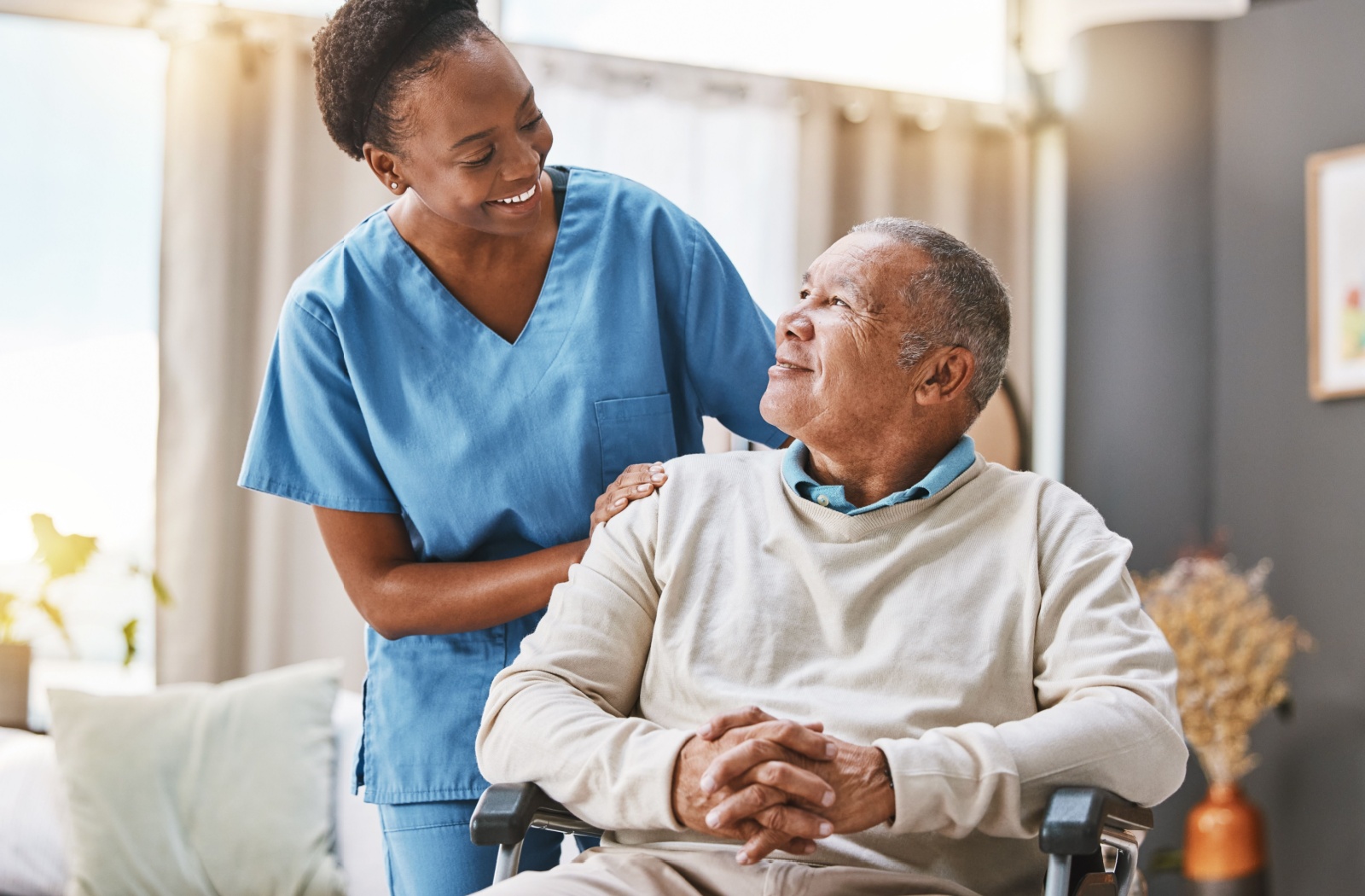 A caregiver smiling warmly at an older adult in a chair, showcasing compassionate support in an assisted living setting.